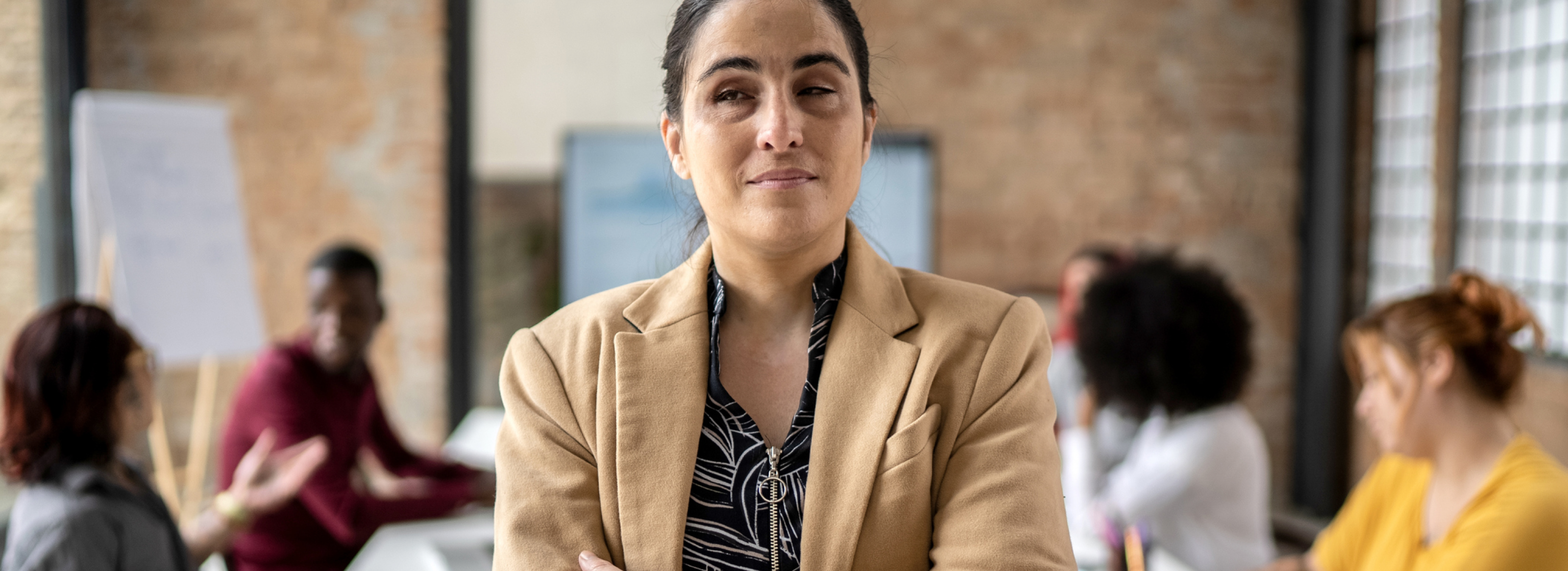 A Visually impaired business woman, stands with confidence. Behind her, a active meeting with colleagues around a table.